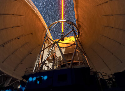 This image shows Gemini North, one half of the International Gemini Observatory, operated by NSF NOIRLab, using a yellow-colored laser beam to adjust its adaptive optics system during the commissioning of its new TOPTICA laser in 2018. This laser emanates from just above the telescope and travels through Earth’s atmosphere to a layer roughly 90 kilometers (50 miles) up. There, it excites traces of sodium gas to produce a glowing ‘artificial star’ which is then used to sample the atmospheric turbulence at lower altitudes. This measurement allows the adaptive optics system to move deformable mirrors on the telescope to compensate for the turbulence. This system allows Gemini North to achieve images of stars, planets, and galaxies at resolutions that rival space-based telescopes.  The laser may seem bright, but this is due to the long exposure of the picture — you can see the stars trailing across the night sky through the open dome indicating the long exposure time. The TOPTICA laser of the observatory is generally dim to the naked eye, depending on how close you are standing to it. You can see another view of the commissioning here.
